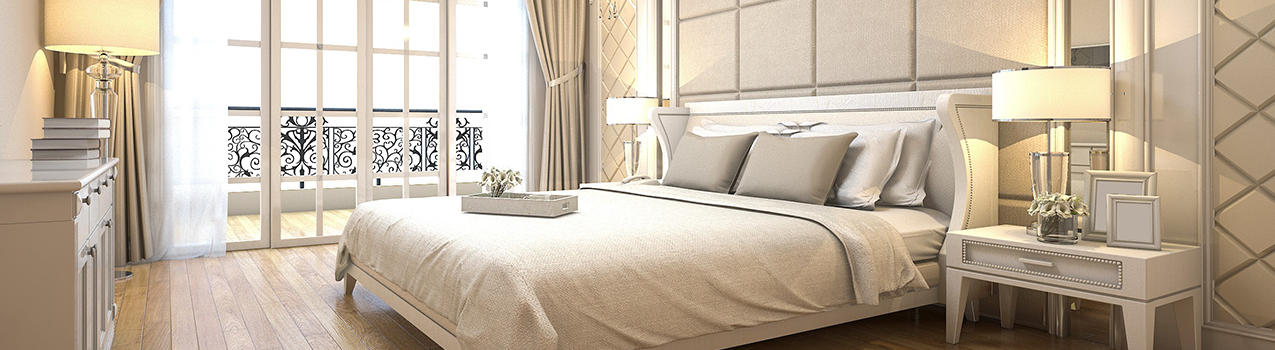 Imagem de quarto em tons claros com janelas no fundo e cama box queen com travesseiros apoiados na cabeceira branca.